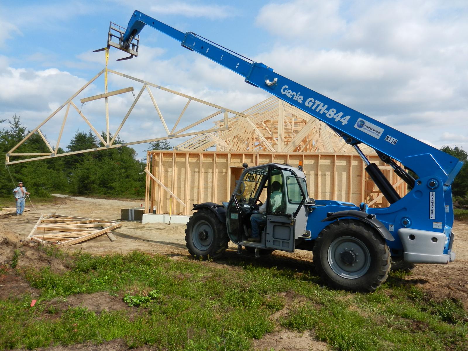 A telehandler lifting a TrussGuy roof truss into place on a framed building